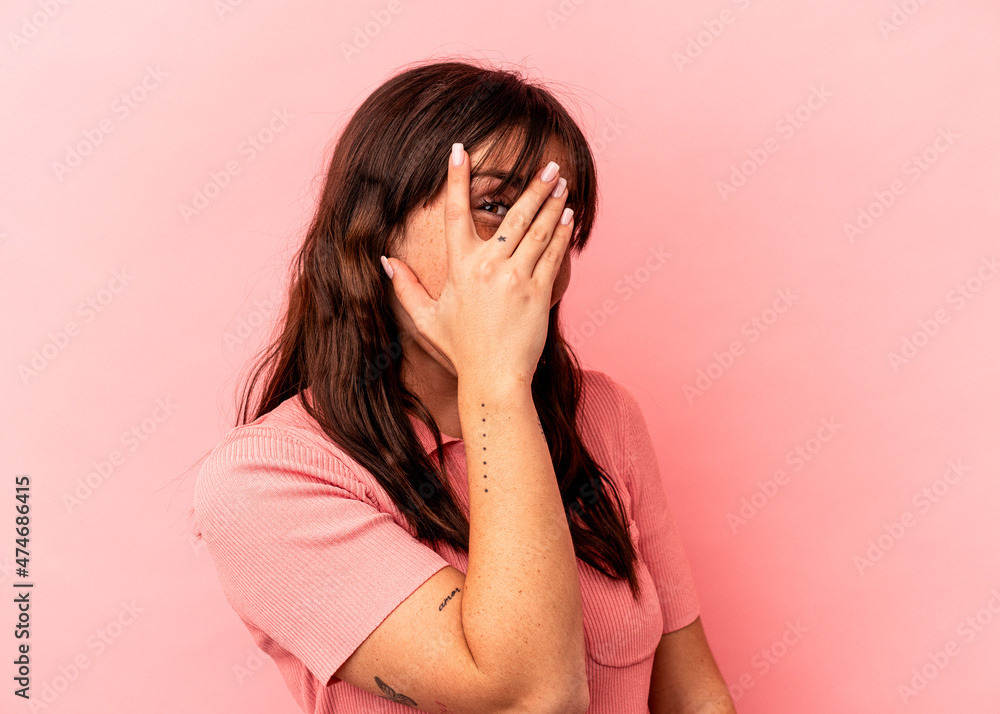 Young Argentinian woman isolated on pink background blink at the camera through fingers, embarrassed covering face. Young Argentinian woman isolated on pink background blink at the camera through fingers, embarrassed covering face.