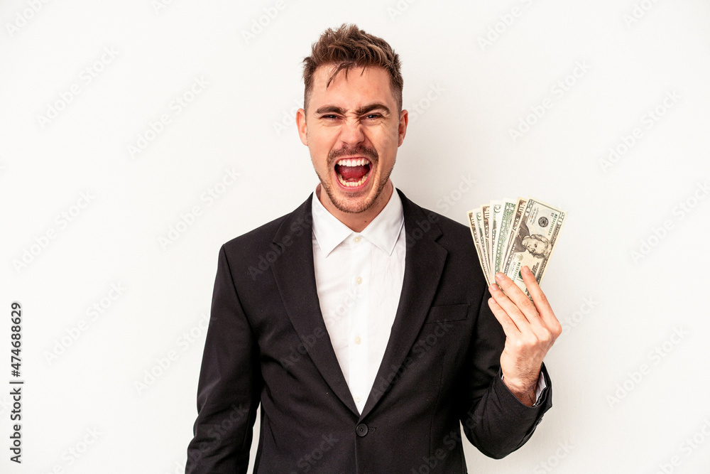 Young caucasian business man holding bank notes isolated on white background screaming very angry and aggressive. Young caucasian business man holding bank notes isolated on white background screaming very angry and aggressive.