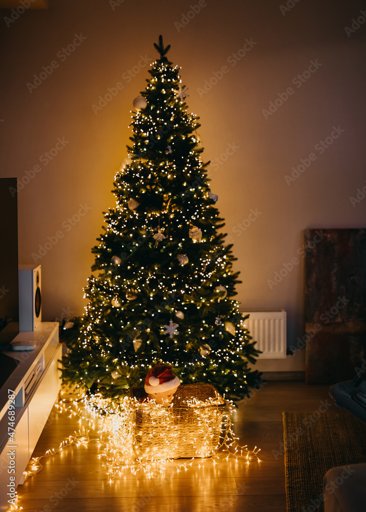 Fototapeta premium Baby in Santa hat, sitting in a basket under a Christmas tree with lights at home, in the dark.