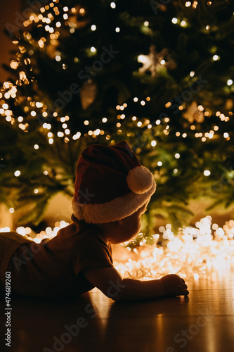 Baby in Santa hat, lying on tummy on the floor, on a Christmas tree with lights background.
