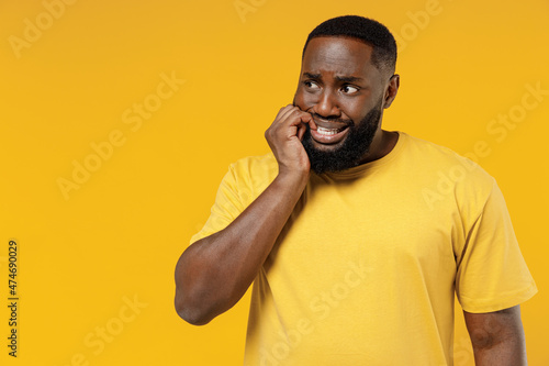 Young puzzled sad black man 20s wear bright casual t-shirt look aside on copy space workspace biting nails fingers isolated on plain yellow color background studio portrait. People lifestyle concept.