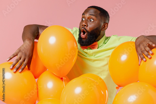 Young shocked astonished black gay man 20s wear green t-shirt bow tie hold bunch of air inflated helium balloons celebrating birthday party isolated on plain pastel pink background studio portrait © ViDi Studio