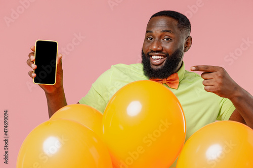 Young black gay man 20s in green t-shirt bow tie hold bunch of air inflated helium balloons holding use mobile cell phone with blank screen workspace area isolated on plain pastel pink background © ViDi Studio
