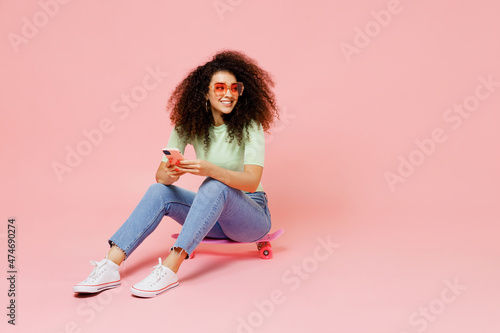 Full size body length young curly latin woman 20s wear casual clothes sunglasses sit on skateboard look aside hold use mobile cell phone isolated on plain pastel light pink background studio portrait