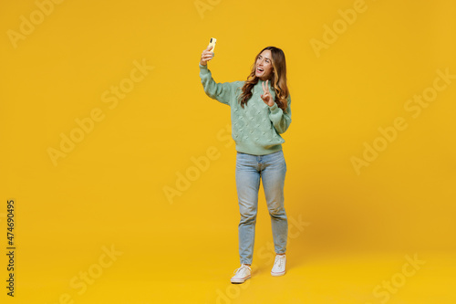 Full body young smiling woman 30s wearing green knitted sweater doing selfie shot on mobile cell phone show v-sign isolated on plain yellow color background studio portrait. People lifestyle concept. © ViDi Studio