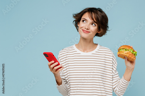 Young minded fun woman in striped shirt using mobile cell phone hold burger count calories browsing internet think isolated on plain pastel light blue background studio People lifestyle food concept photo