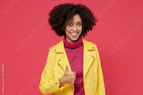 Charming bright happy young curly black latin woman 20s years old wears yellow jacket showing thumb up like gesture isolated on plain red background studio portrait. People emotions lifestyle concept.