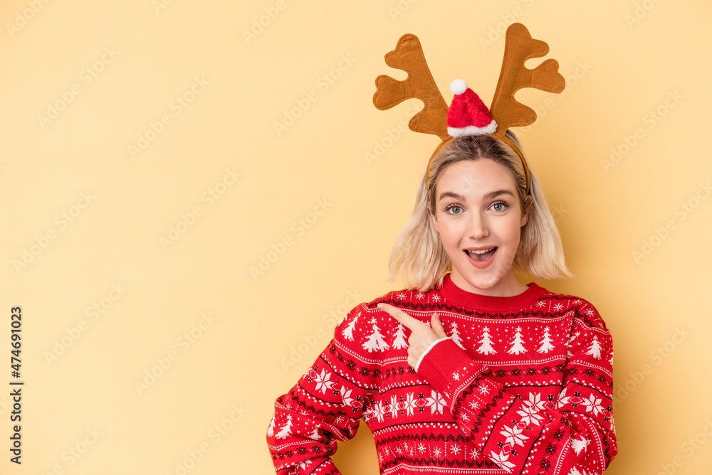 Young caucasian woman wearing a christmas reindeer hat isolated on yellow background smiling and pointing aside, showing something at blank space. Young caucasian woman wearing a christmas reindeer hat isolated on yellow background smiling and pointing aside, showing something at blank space.