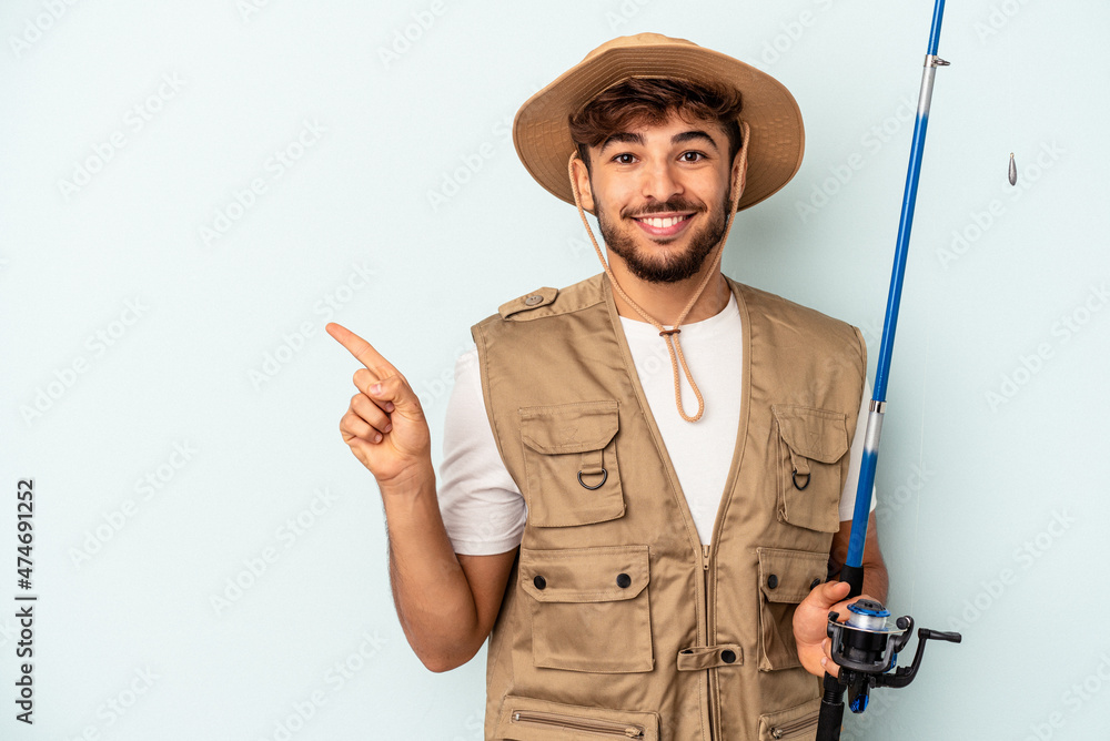 Young mixed race fisherman holding a rod isolated on blue background smiling and pointing aside, showing something at blank space. Young mixed race fisherman holding a rod isolated on blue background smiling and pointing aside, showing something at blank space.