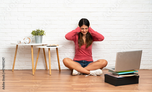 Young woman with a laptop sitting on the floor at indoors frustrated and covering ears