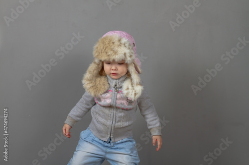 Cute baby girl 2 years old caucasian in warm winter clothes and a hat with earflaps with fur on a gray background