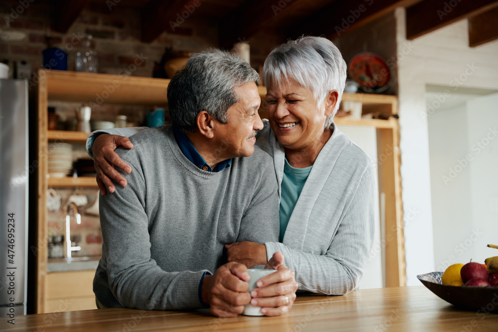 Happily retired elderly biracial couple smiling at each other. Wife holding husband in modern kitchen. Happily retired elderly biracial couple smiling at each other. Wife holding husband in modern kitchen.