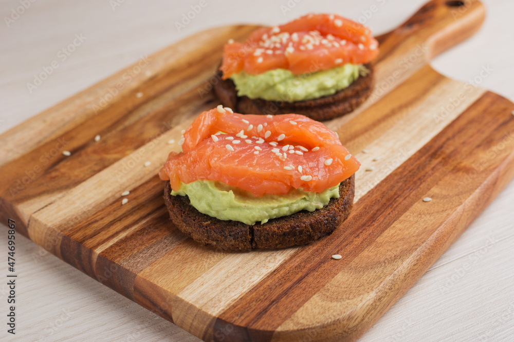 Close up of toasty bread with avocado, salmon, sesame seeds served on wooden board. Healthy breakfast. Selective focus.