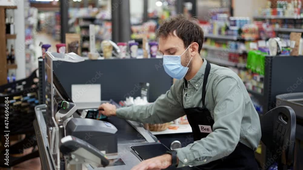 Sales clerk in mask working at grocery store. Male cashier in black apron at cash desk and unrecognizable customer Sales clerk in mask working at grocery store. Male cashier in black apron at cash desk and unrecognizable customer