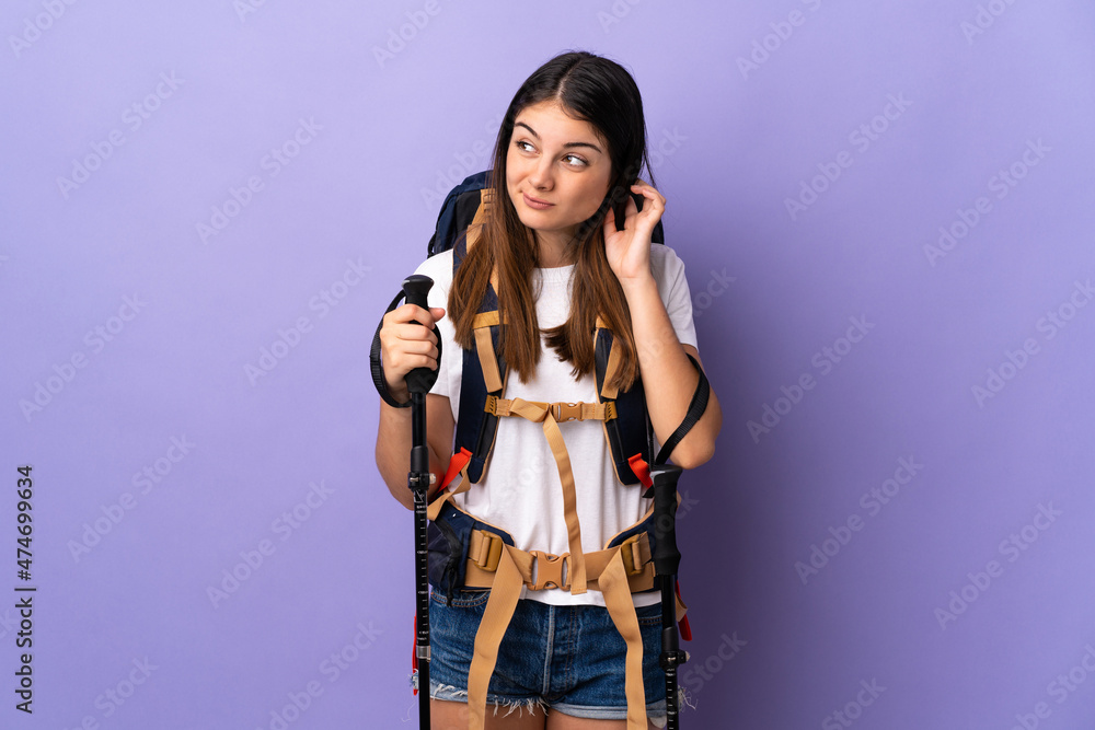 Young woman with backpack and trekking poles isolated on purple background having doubts