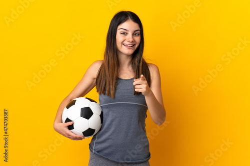 Young football player woman isolated on yellow background surprised and pointing front © luismolinero