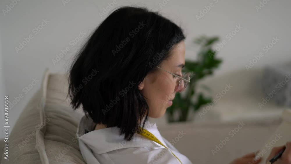 Cropped image of fashion designer draws cloth sketch in notebook. Female creating new design while sitting with a centimeter on the shoulders near open laptop computer. Student learning on sketching