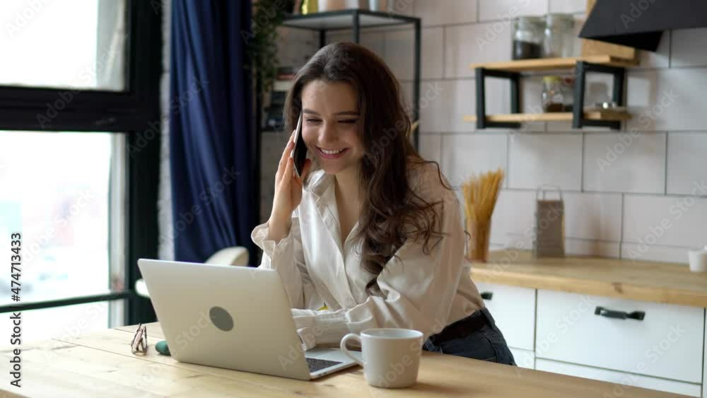 Young female having mobile phone conversation while sitting front open laptop computer at home kitchen interior. Attractive brunette woman calling with smartphone for discuss work issues Young female having mobile phone conversation while sitting front open laptop computer at home kitchen interior. Attractive brunette woman calling with smartphone for discuss work issues