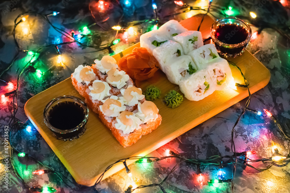 Appetizing and delicious sushi rolls with shrimp, cheese, caviar, chicken, soy sauce, nori, wasabi and ginger on a wooden board on a dark background with a Christmas garland