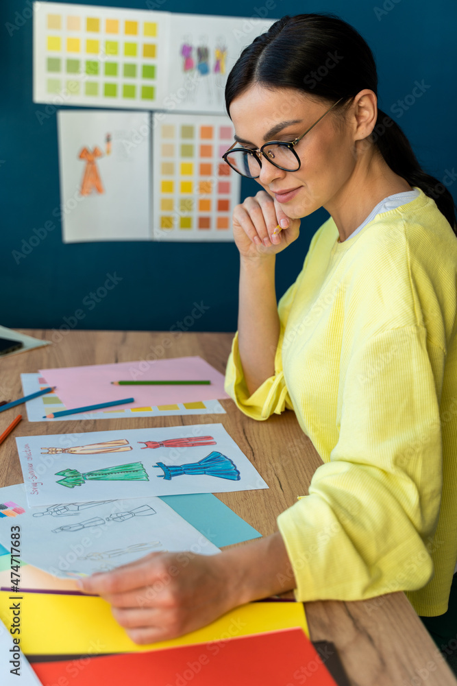 Vertical view of pensive brunette couturier using pencil for modeling sketches while drawing on paper in showroom. Stock photo Vertical view of pensive brunette couturier using pencil for modeling sketches while drawing on paper in showroom. Stock photo