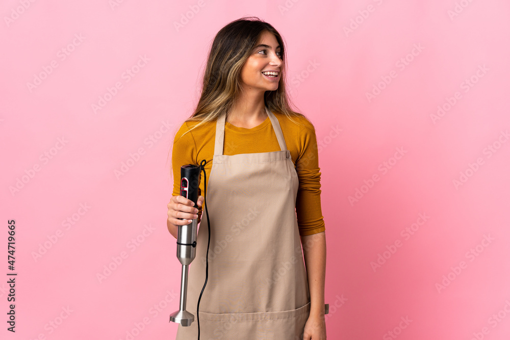 Young chef woman using hand blender isolated on pink background looking side