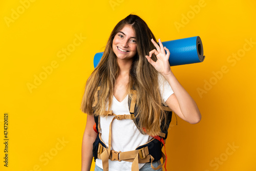 Young mountaineer woman with a big backpack isolated on yellow background showing ok sign with fingers