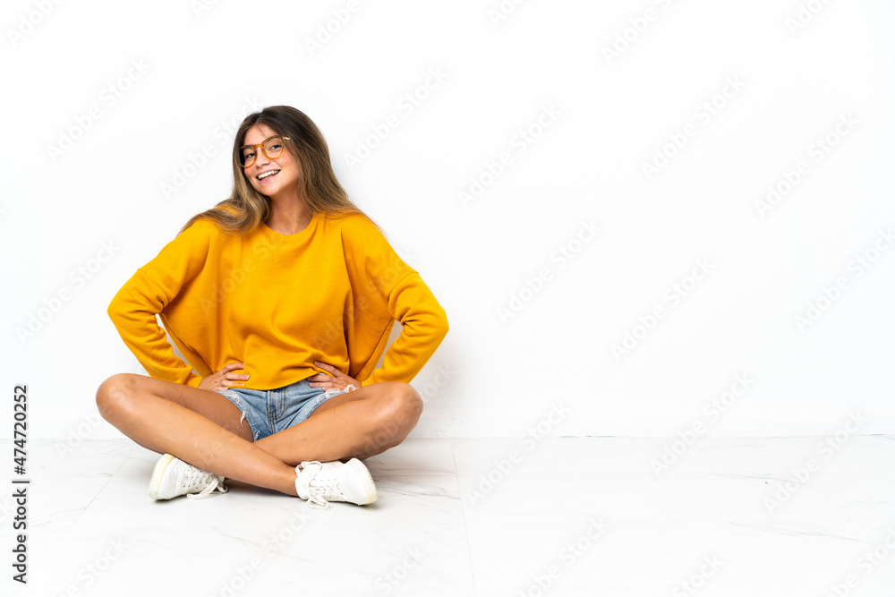 Young woman sitting on the floor isolated on white background posing with arms at hip and smiling