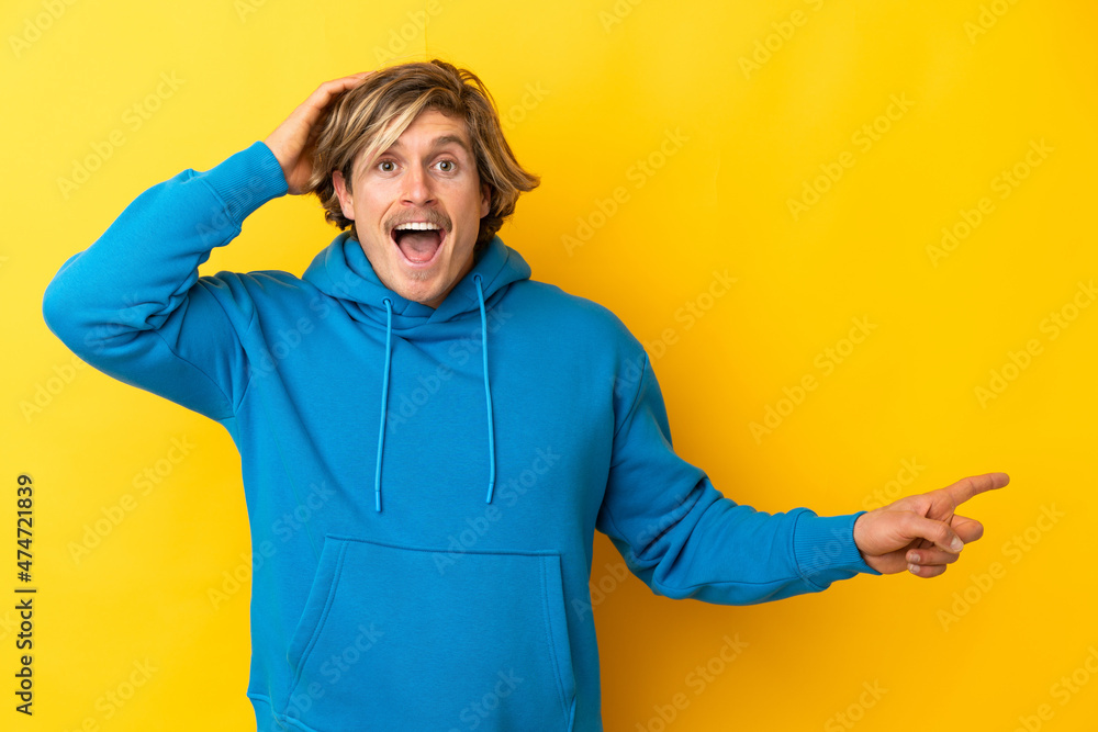 Handsome blonde man isolated on yellow background surprised and pointing finger to the side Handsome blonde man isolated on yellow background surprised and pointing finger to the side