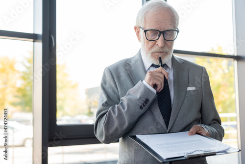 thoughtful senior businessman holding pen while reading contract in office