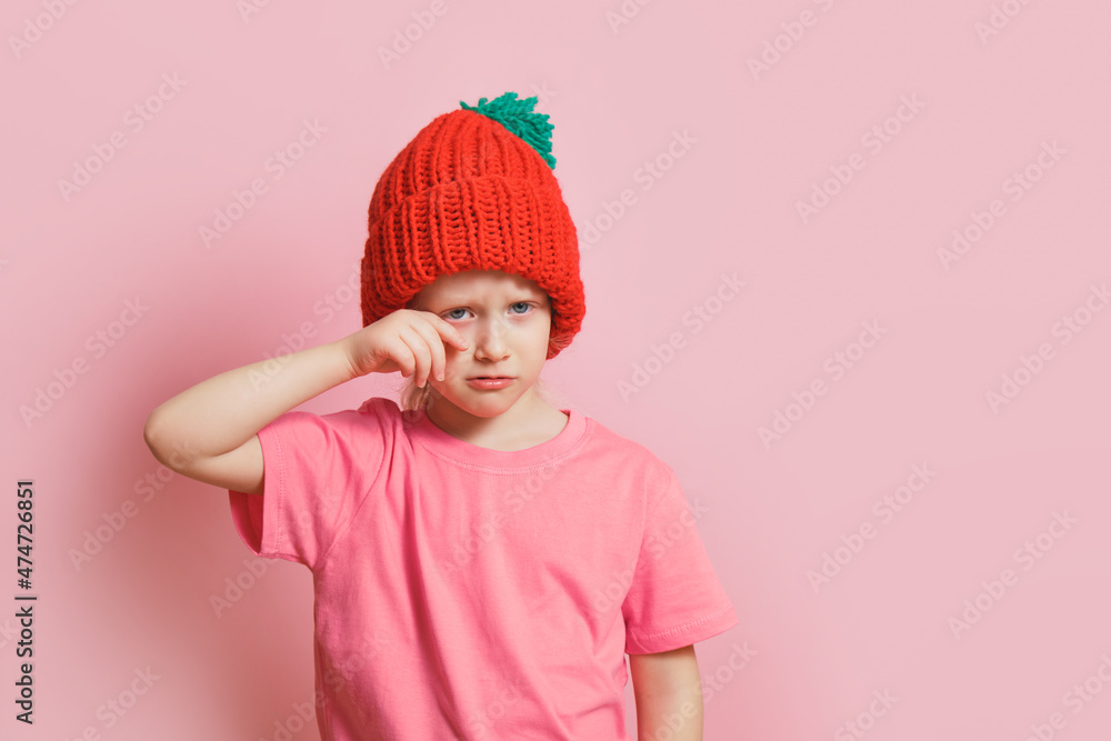 cute caucasian toddler girl in a knitted hat on a pink background, a cowardly child wipes tears cute caucasian toddler girl in a knitted hat on a pink background, a cowardly child wipes tears