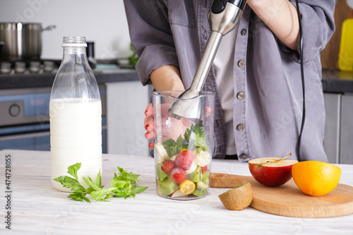Woman using hand blender to make smoothies. Grey kitchen interior design. Vegetarian, clean eating lifestyle concept.