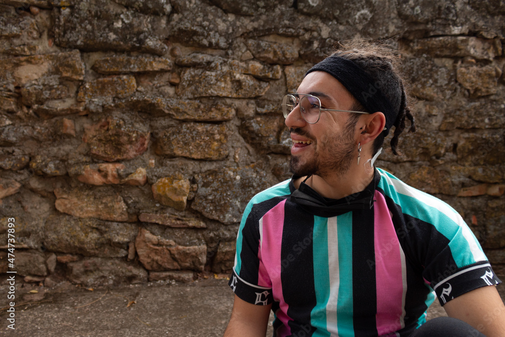 young man with glasses beard and braids in hair and earring smiling happy and looking to the side, stone wall background