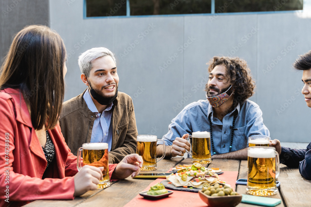 Multiracial people having fun drinking beer at brewery bar outdoor while wearing safety masks - Focus on left guy face