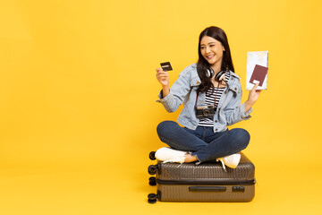 Happy young Asian woman tourist sitting on baggage and holding credit card, maps and passport isolated on yellow background