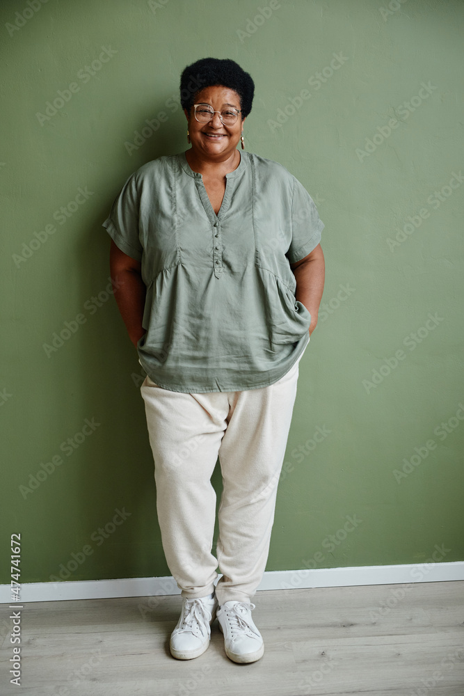 Vertical full length portrait of black senior woman wearing glasses and smiling at camera while standing against green background Vertical full length portrait of black senior woman wearing glasses and smiling at camera while standing against green background