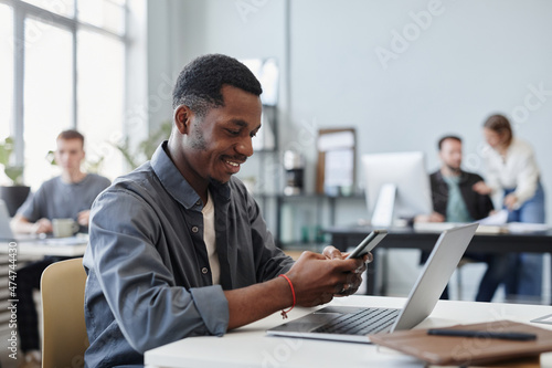 African young businessman typing a message on mobile phone and smiling while sitting at his workplace with laptop at office