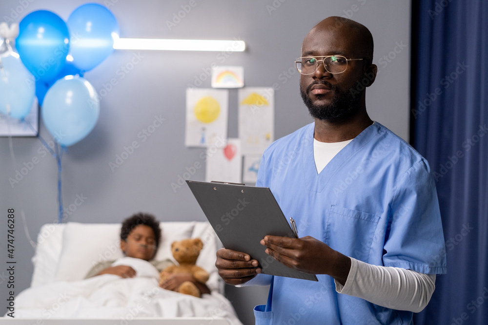 Portrait of African doctor in uniform holding medical card looking at camera while standing at hospital ward with little boy in the background Portrait of African doctor in uniform holding medical card looking at camera while standing at hospital ward with little boy in the background