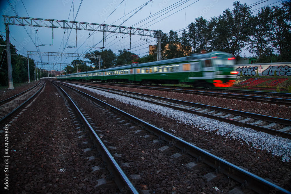 Electric passenger train (motion-blurred) run in summer dusk on Kursk railroad. Blue sky, green trees. Graffiti on fence.