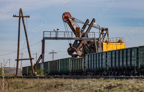 Ore loading station. Yellow excavator loads open wagons. Worker's bridge. Old wood electric pole without wires. Blue sky, clouds. Khromtau, Kazakhstan.