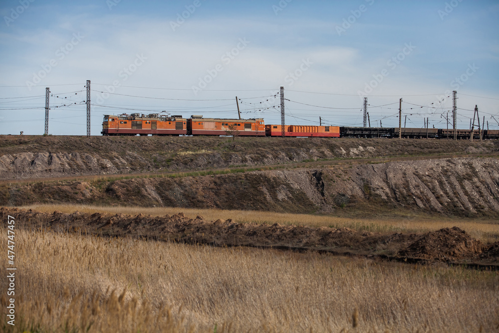 Train with orange locomotive and wagon in steppe. Platform cars for coal and ore transportation. Yellow grass, brown ground. Electric towers, wires, blue sky with cloud. Sunset view. Train with orange locomotive and wagon in steppe. Platform cars for coal and ore transportation. Yellow grass, brown ground. Electric towers, wires, blue sky with cloud. Sunset view.