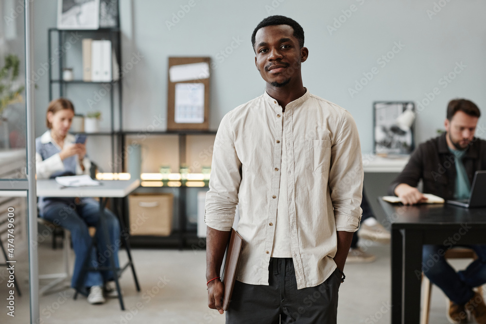 Portrait of African young businessman looking at camera while standing at office with his colleagues in the background Portrait of African young businessman looking at camera while standing at office with his colleagues in the background