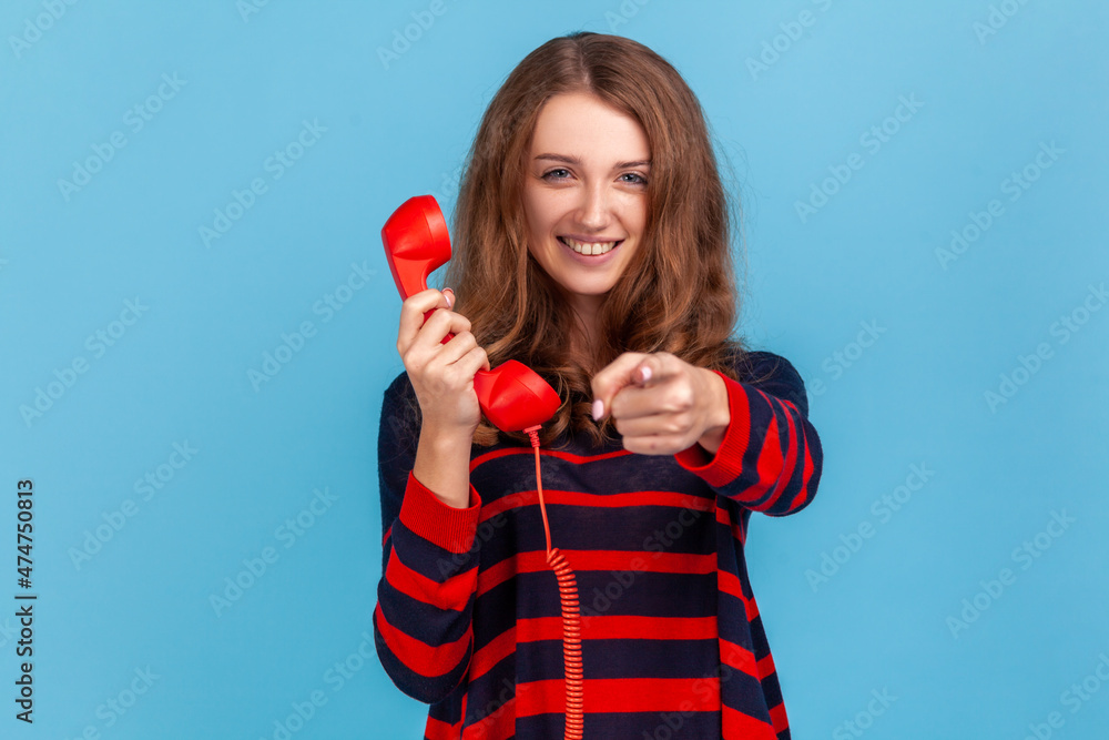 Optimistic woman wearing striped casual style sweater, holding red landline phone and pointing to camera with finger, call me, I am waiting. Indoor studio shot isolated on blue background. Optimistic woman wearing striped casual style sweater, holding red landline phone and pointing to camera with finger, call me, I am waiting. Indoor studio shot isolated on blue background.