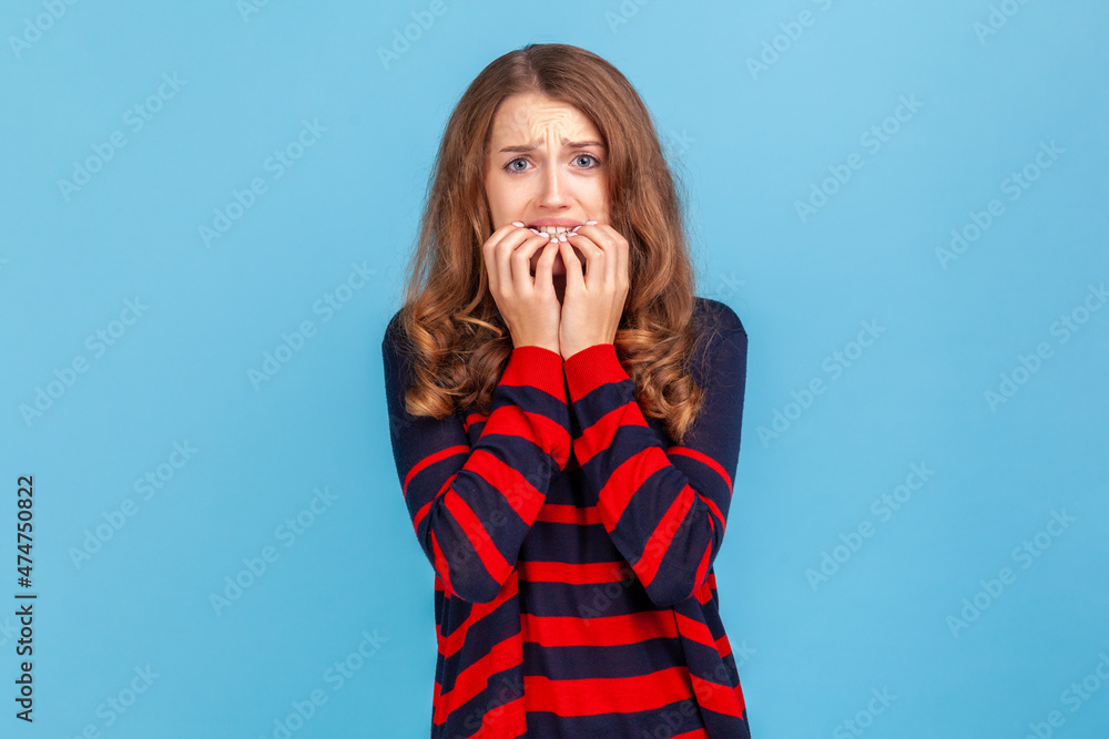 Troubles and worries. Portrait of nervous woman wearing striped sweater biting nails, terrified about problems, suffering phobia, anxiety disorder. Indoor studio shot isolated on blue background. Troubles and worries. Portrait of nervous woman wearing striped sweater biting nails, terrified about problems, suffering phobia, anxiety disorder. Indoor studio shot isolated on blue background.