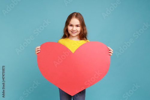 Portrait of charming little girl holding in hands large red heart symbol, looking smiling at camera, wearing yellow casual style sweater. Indoor studio shot isolated on blue background.
