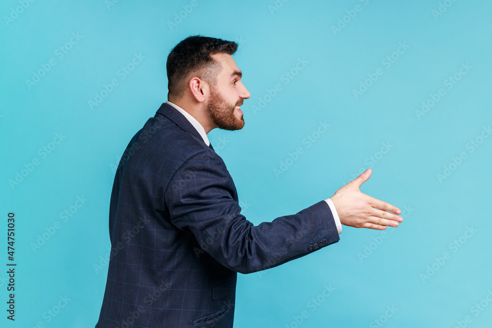 Hi, nice to meet you! Side view of friendly smiling handsome man wearing official style suit outstretching hand, offering handshake on job interview. Indoor studio shot isolated on blue background. Hi, nice to meet you! Side view of friendly smiling handsome man wearing official style suit outstretching hand, offering handshake on job interview. Indoor studio shot isolated on blue background.