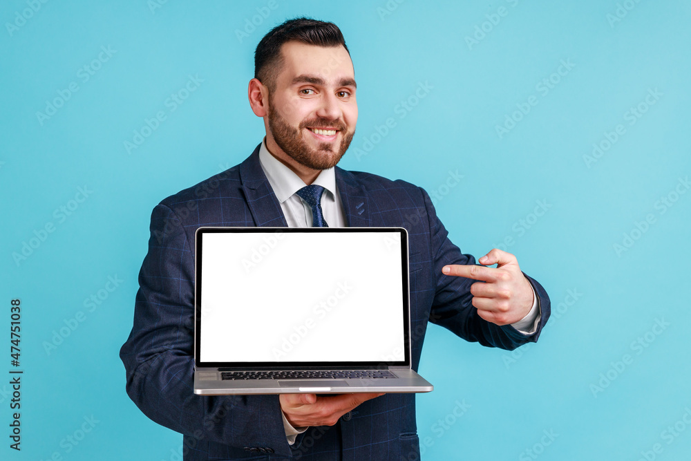 Portrait of positive joyful businessman in official style suit standing holding laptop with blank screen and smiling at camera, internet advertising. Indoor studio shot isolated on blue background. Portrait of positive joyful businessman in official style suit standing holding laptop with blank screen and smiling at camera, internet advertising. Indoor studio shot isolated on blue background.