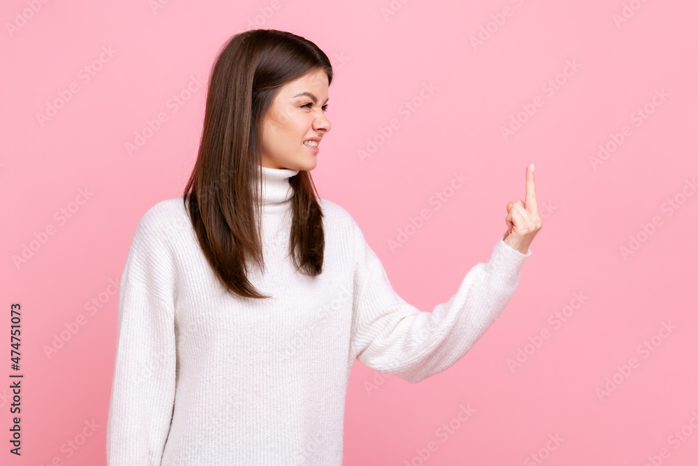 Side view portrait of aggressive woman standing and shows fuck sign, expressing negative emotions, wearing white casual style sweater. Indoor studio shot isolated on pink background. Side view portrait of aggressive woman standing and shows fuck sign, expressing negative emotions, wearing white casual style sweater. Indoor studio shot isolated on pink background.