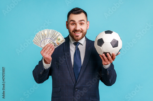 Obraz na plátně Positive man wearing official style suit holding soccer ball and hundred dollar bills, looking camera with smile , betting and winning