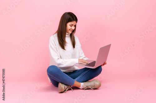Portrait of happy brunette girl sitting on floor with crossed legs and working on laptop, freelancer, wearing white casual style sweater. Indoor studio shot isolated on pink background.