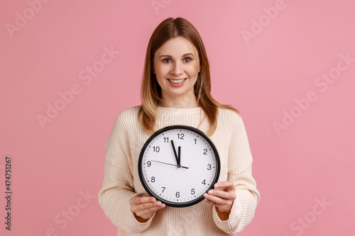 Portrait of happy positive blond woman holding big wall clock, looking at camera with pleasant smile, time to go, wearing white sweater. Indoor studio shot isolated on pink background.